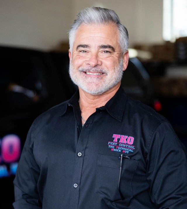Smiling man in a black shirt with a logo, standing indoors against a blurred dark background.