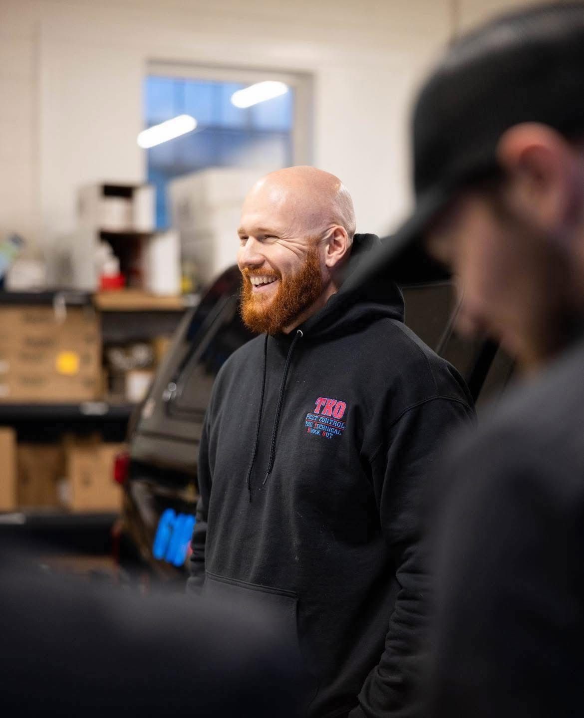 Smiling red-bearded man in a black hoodie standing in a workshop with blurred colleagues in the foreground