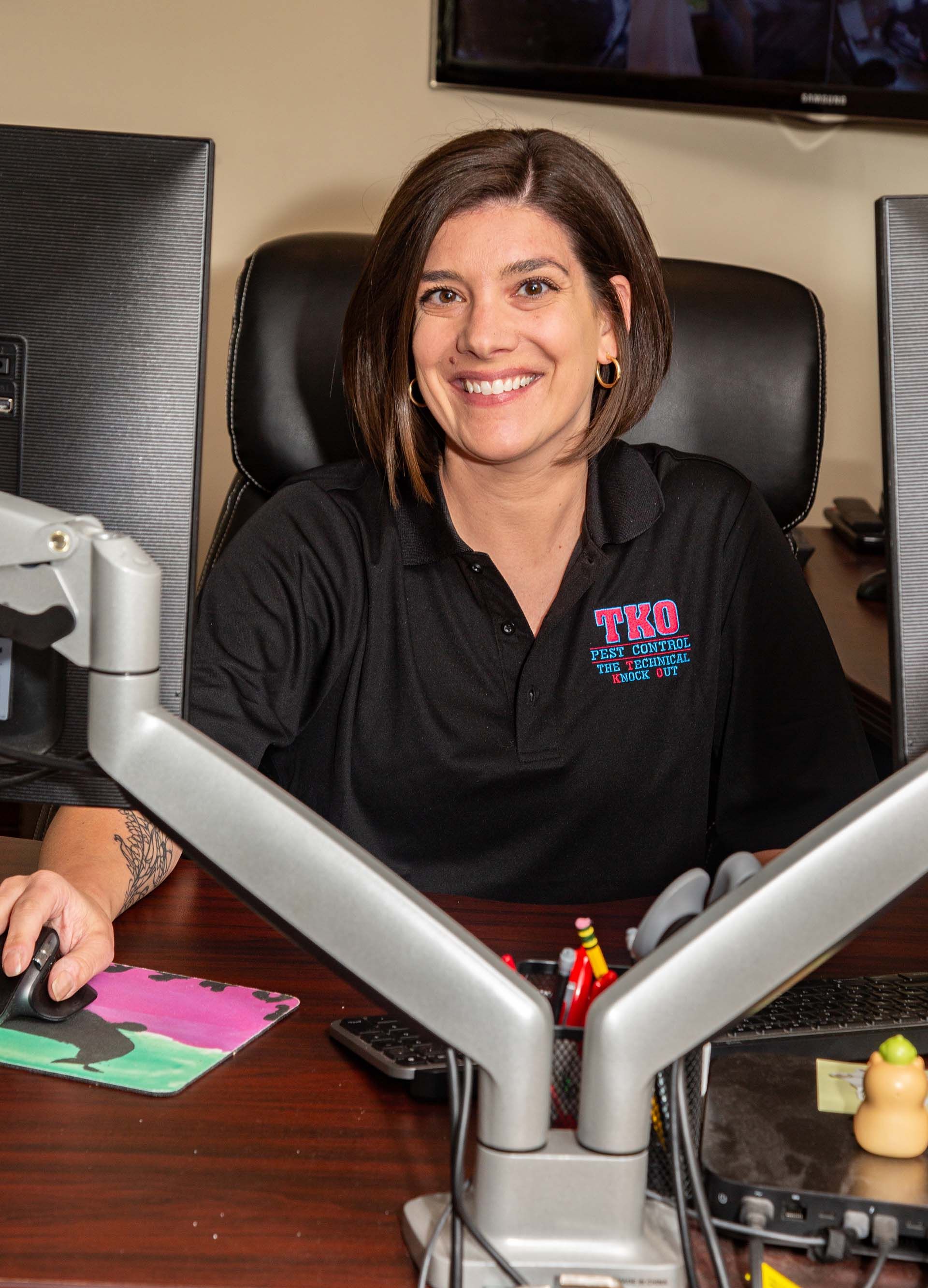 Woman smiling at a desk in a black polo, seated in front of a microphone in a studio or office setting
