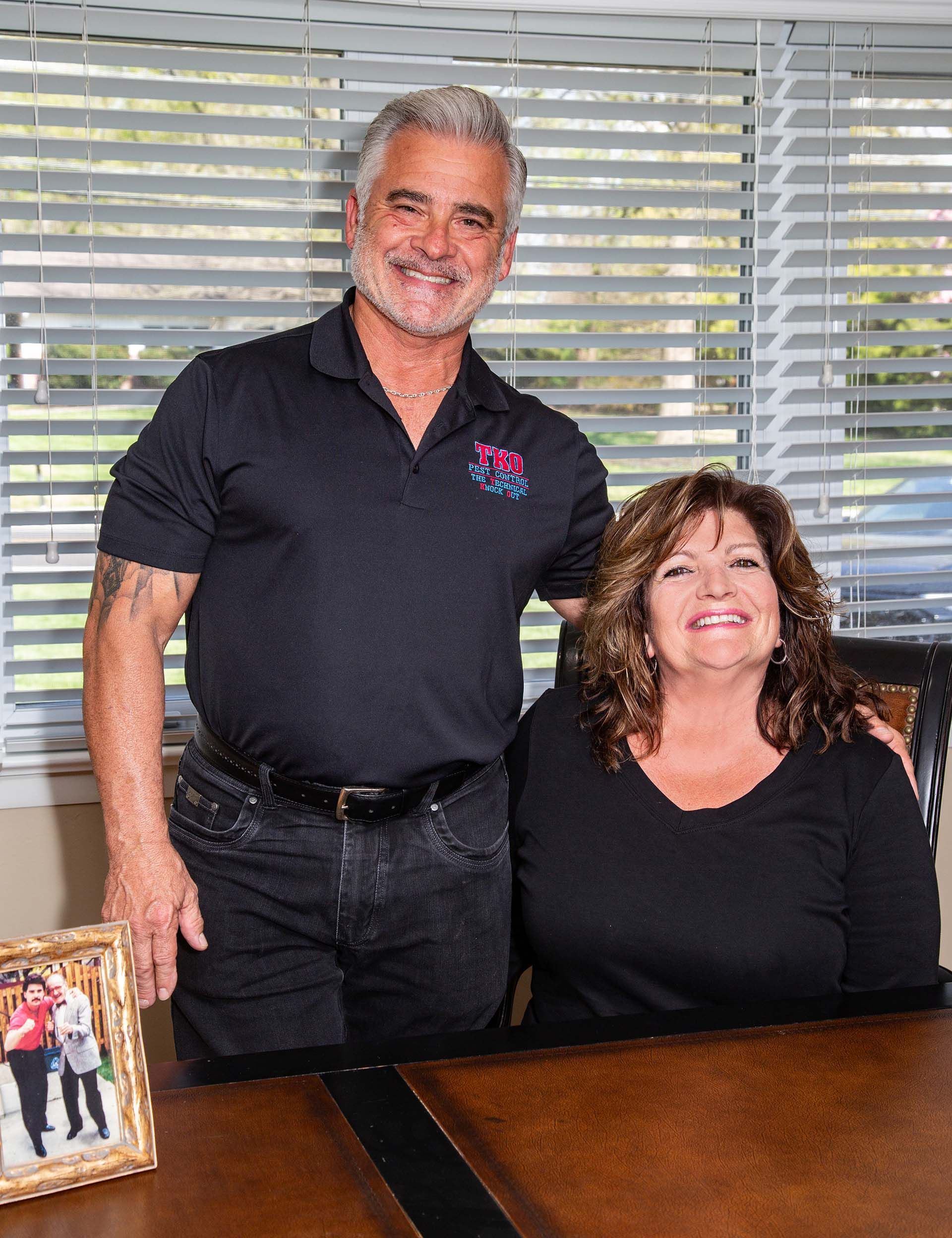 Two people posing indoors beside a desk, smiling in front of window blinds.