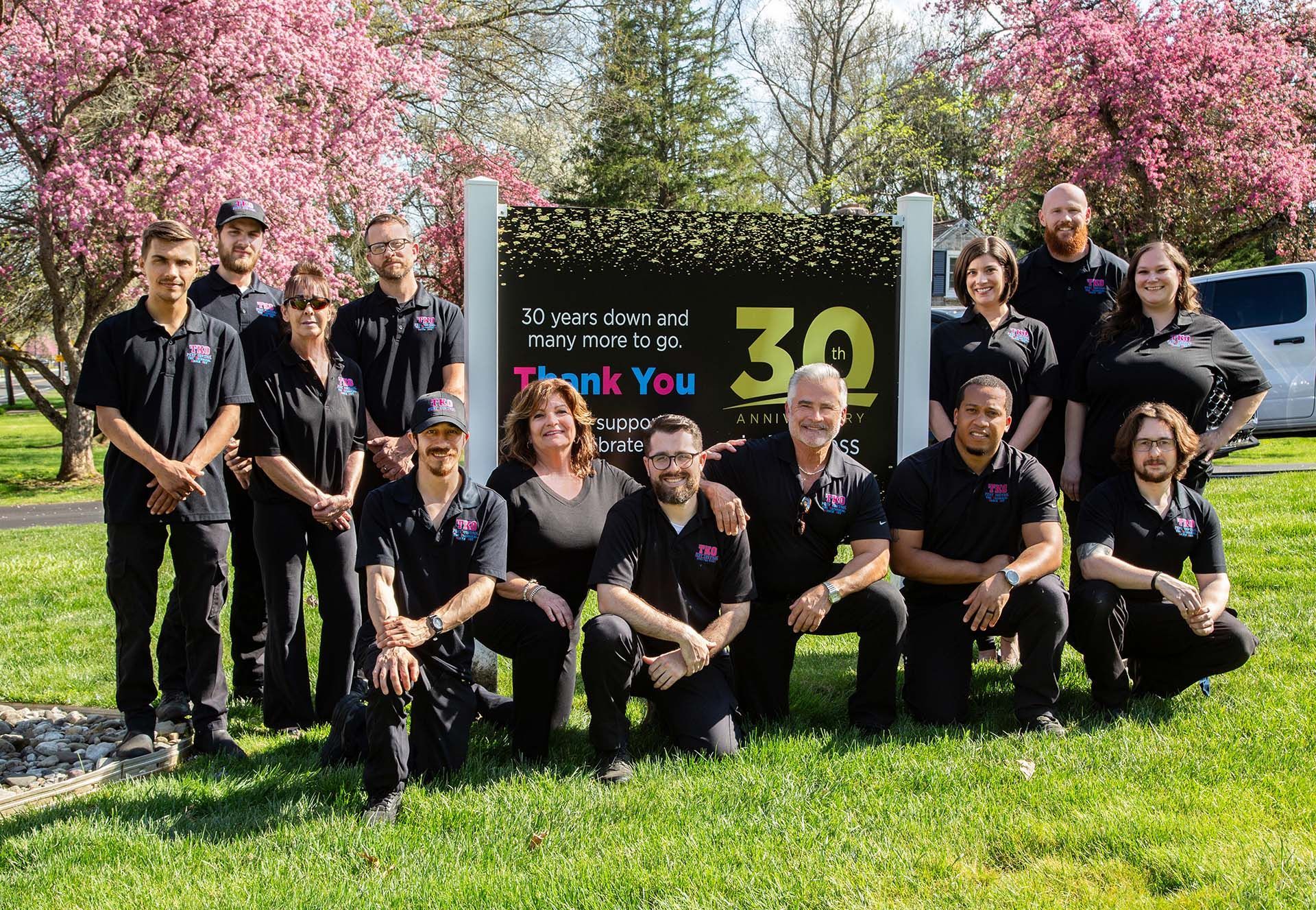 Group posing outdoors in front of a sign with “30” and “Thank You,” with pink flowering trees behind them