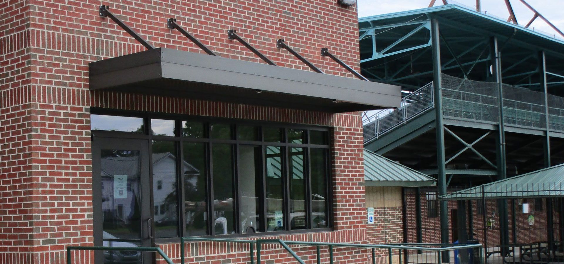 Brick storefront entrance with brown awning and large windows beside a metal stadium structure