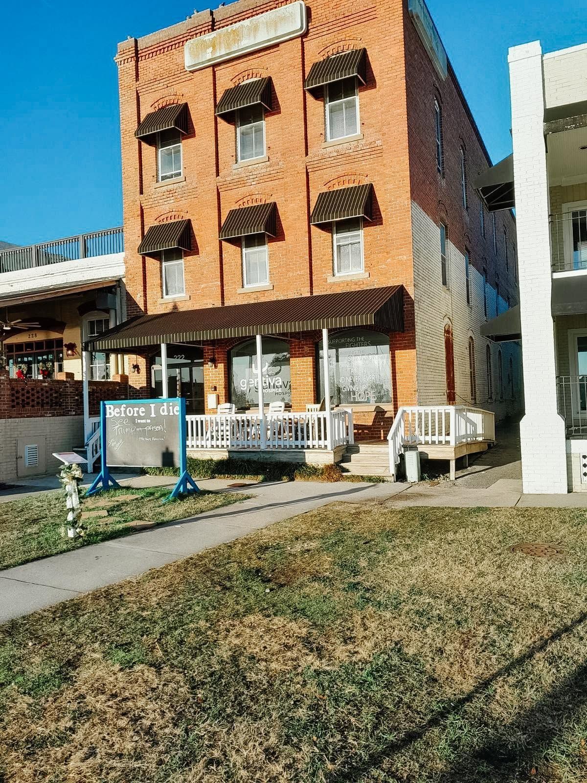 Three-story brick building with striped awnings, benches, and a small lawn in front