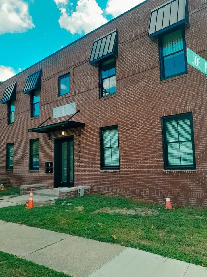 Brick apartment building with black awnings, green lawn, and a sidewalk under a partly cloudy sky