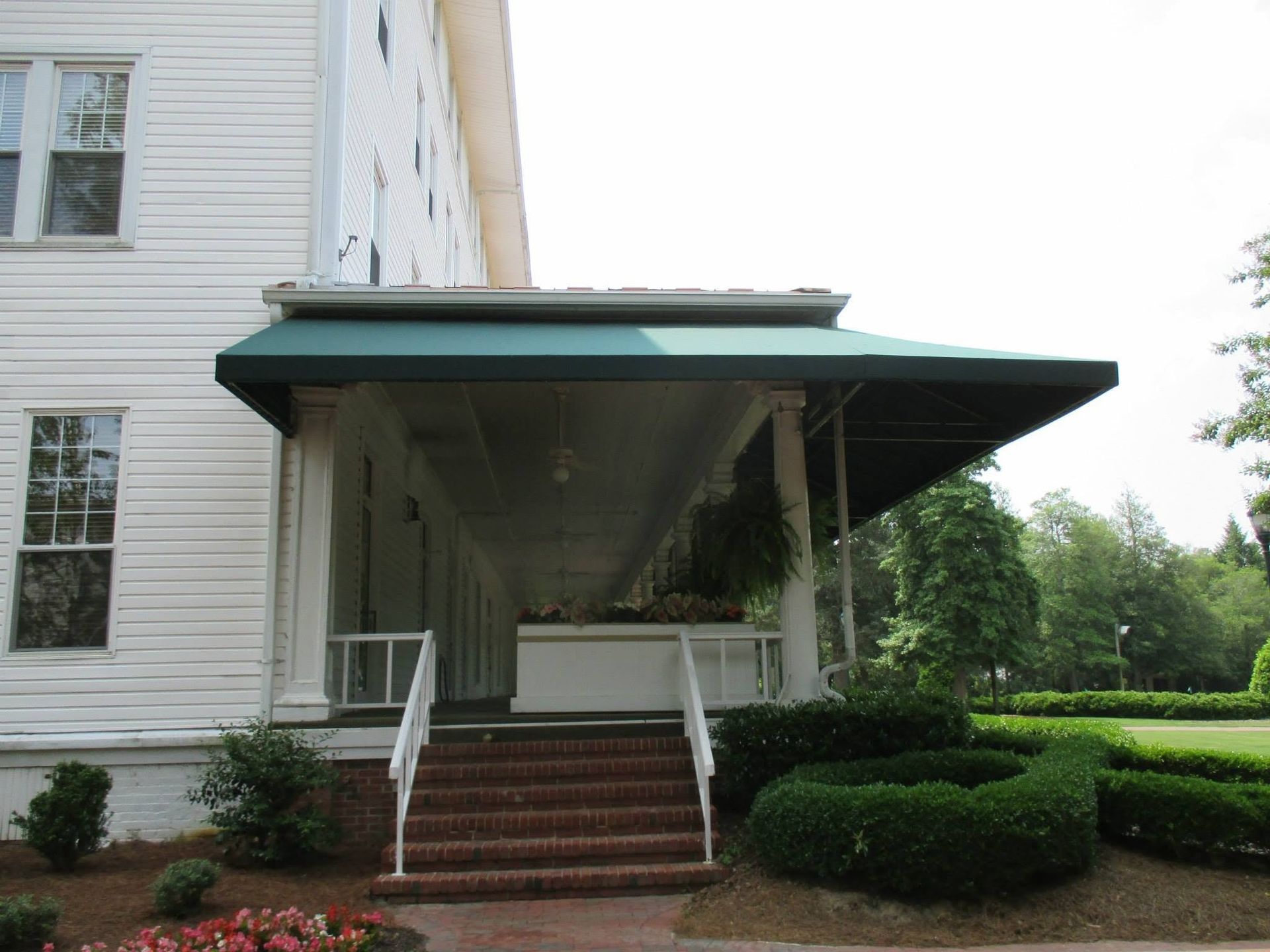 Covered porch with green awning and brick steps on a white house, with shrubs along the front walk