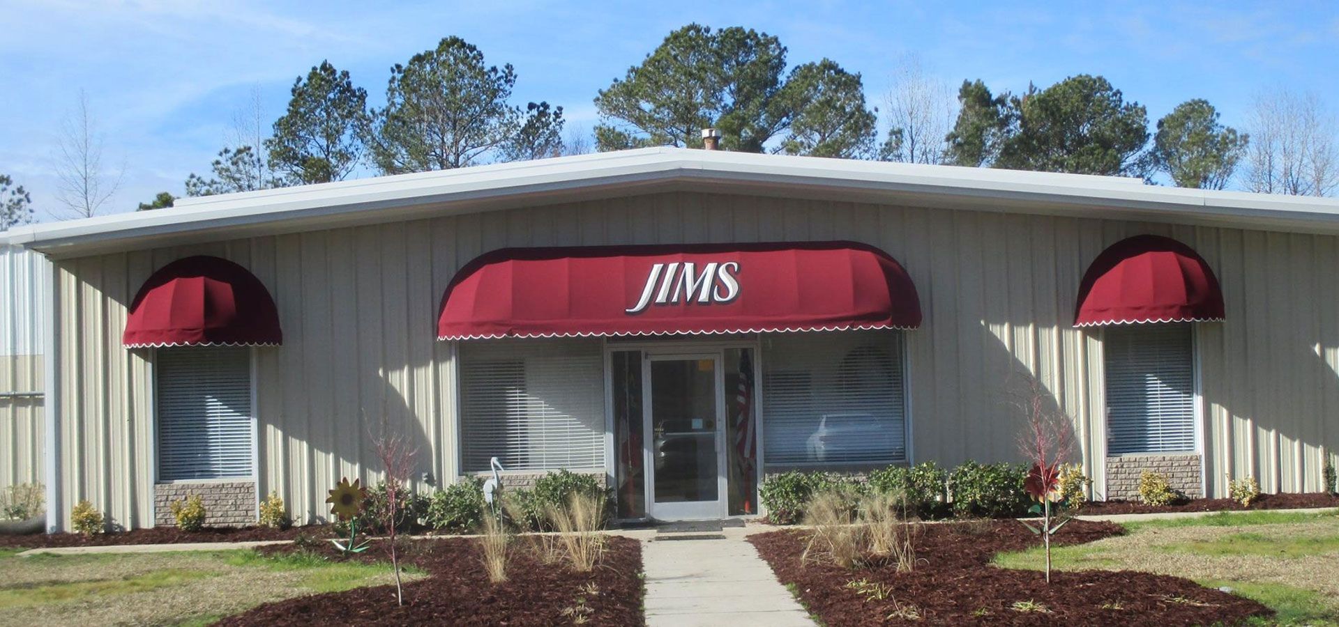 Front view of a gray building with red awnings and a central entrance labeled “JIMS.”