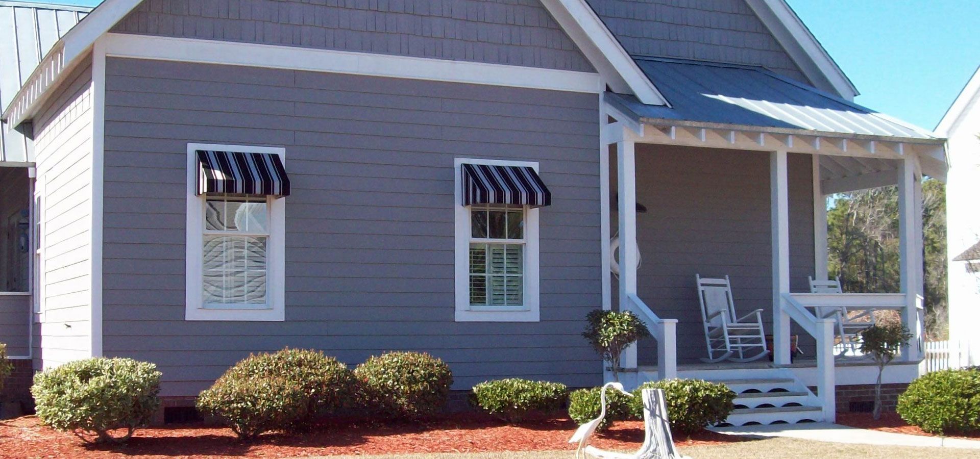 Small gray house with striped awnings, white porch, and landscaped shrubs in front yard