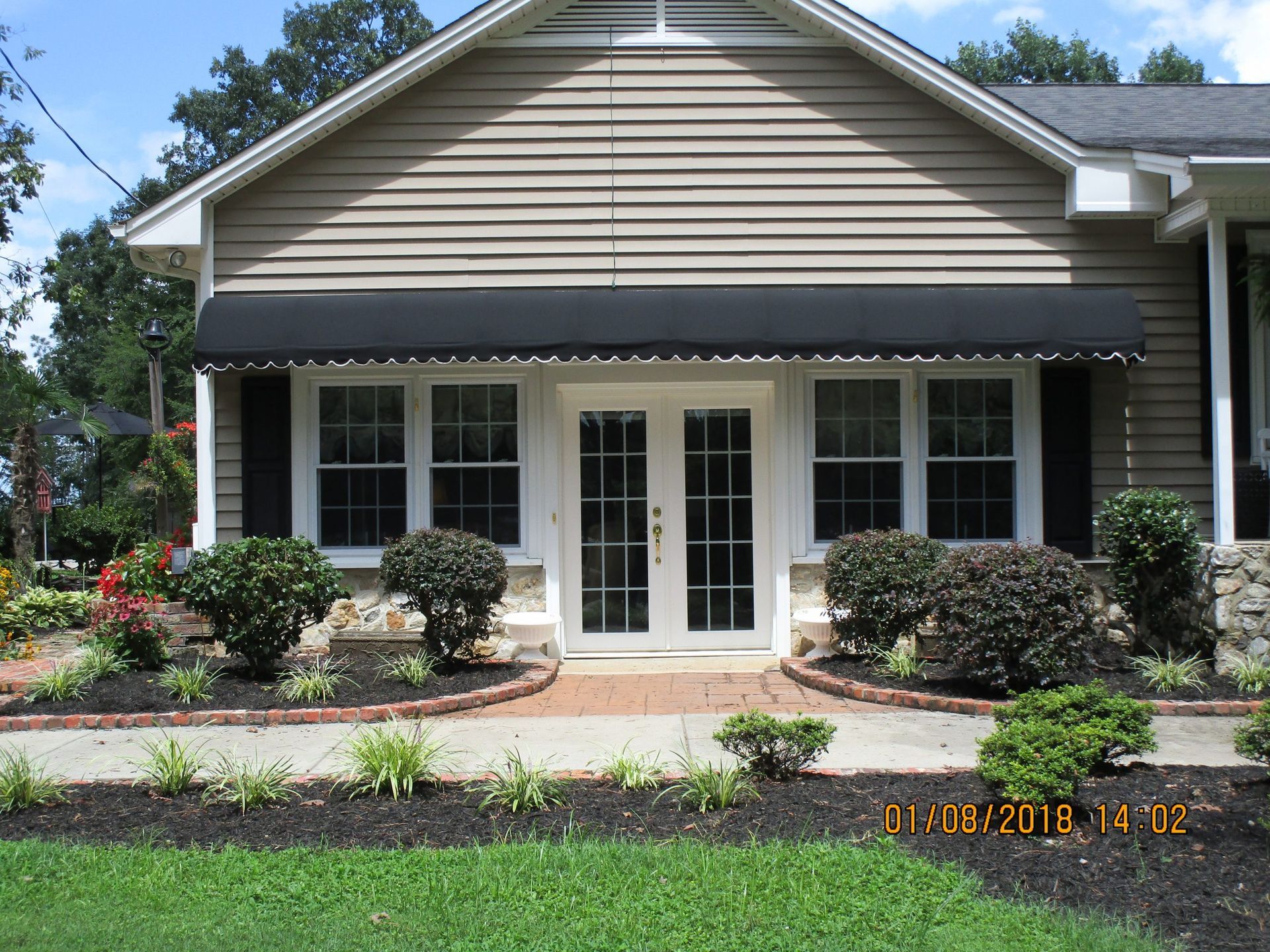 Suburban house exterior with white French doors, black awning, and landscaped flower beds