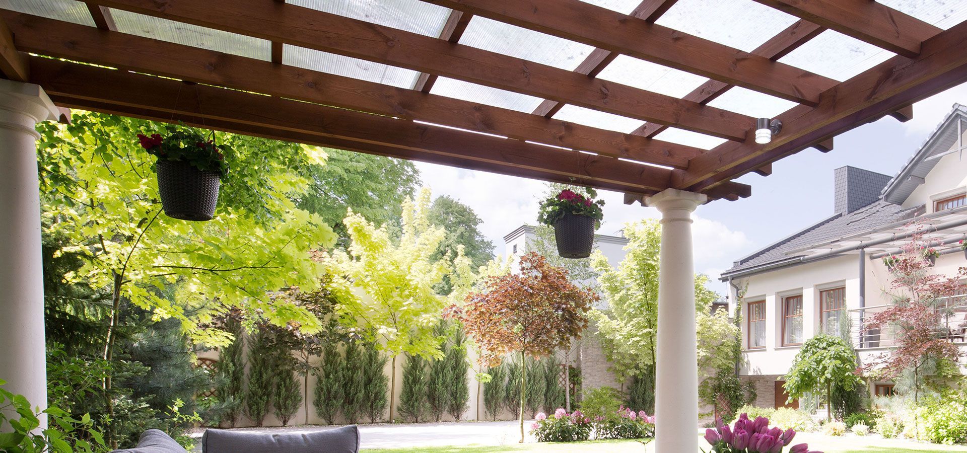 Sunny garden patio with wooden pergola, hanging plants, and a white house in the background