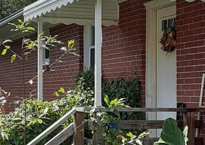 Red brick house porch with white trim, plants, a front door wreath, and a staircase railing in the foreground