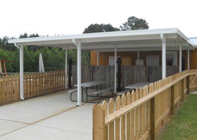 Covered porch with picnic tables and a wooden fence, leading to a ramp at a park pavilion