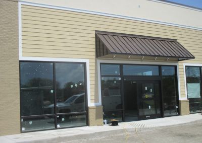 Empty storefront with tan siding, dark windows, and a brown awning at a strip mall.