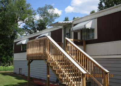 Mobile home with wooden stairs and railing, front awning, and trees in the yard.