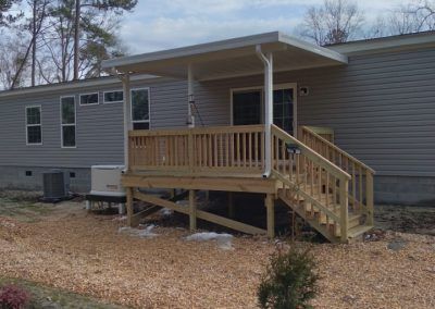 Mobile home with a small wooden front porch and stairs on a gravel lot, with trees in the background