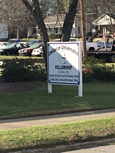 University sign for a fellowship program on a grassy roadside near parked cars and buildings.