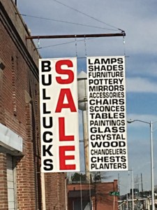 Street sign for a store selling bullucks, sale, lamps, shades, furniture, mirrors, accessories, chairs, and plants.