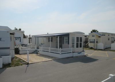Small white mobile home with porch in a trailer park, beside a paved road under a clear sky