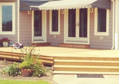 Front porch with beige siding, striped awnings, steps, and potted plants beside a patio bench.