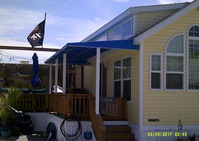 Yellow house with porch and blue awnings, black flag, and patio furniture in sunlight