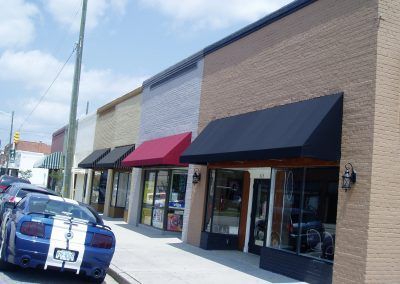 Row of storefronts with black and red awnings beside a parked blue car on a sunny street