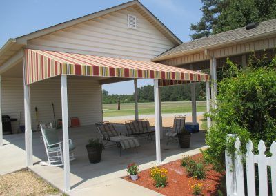 Striped awning shading a porch with rocking chairs beside a white house and picket fence