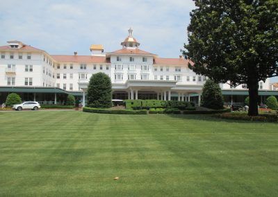 Large white hotel with red roofs behind a wide green lawn and trees
