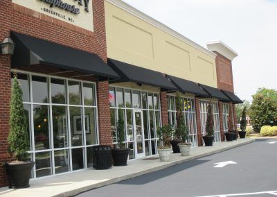 Storefronts with black awnings and large glass windows along a shopping plaza parking lot