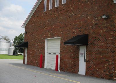 Brick building exterior with white doors, black awnings, and red bollards by a paved lot