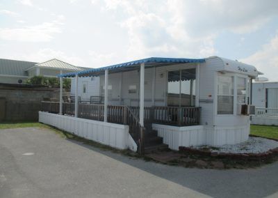 White mobile home with blue roof on a paved lot, with steps, porch railings, and covered front deck.
