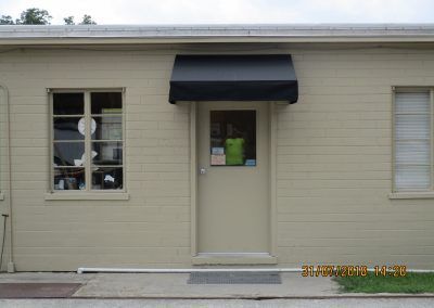 Front of a beige building with a black awning over the center door and windows on both sides.
