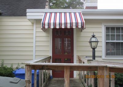 Front porch with red door, striped awning, wooden ramp, and black lantern by a house entrance