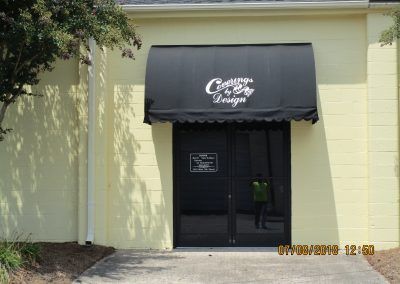Black awning over a glass entrance on a pale yellow building, with a small sign and trees nearby