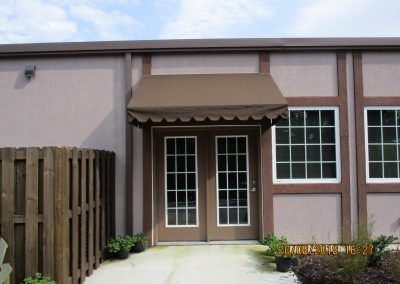 Front entrance of a beige building with a brown awning, glass doors, and flower beds along the walkway