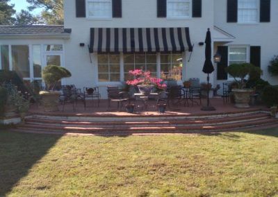 Front of a white house with black-striped awning, brick steps, patio table, and lawn in front