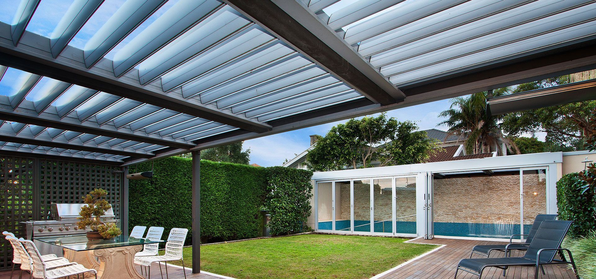Covered patio with dining table, lounge chairs, and a backyard pool beside a hedge wall