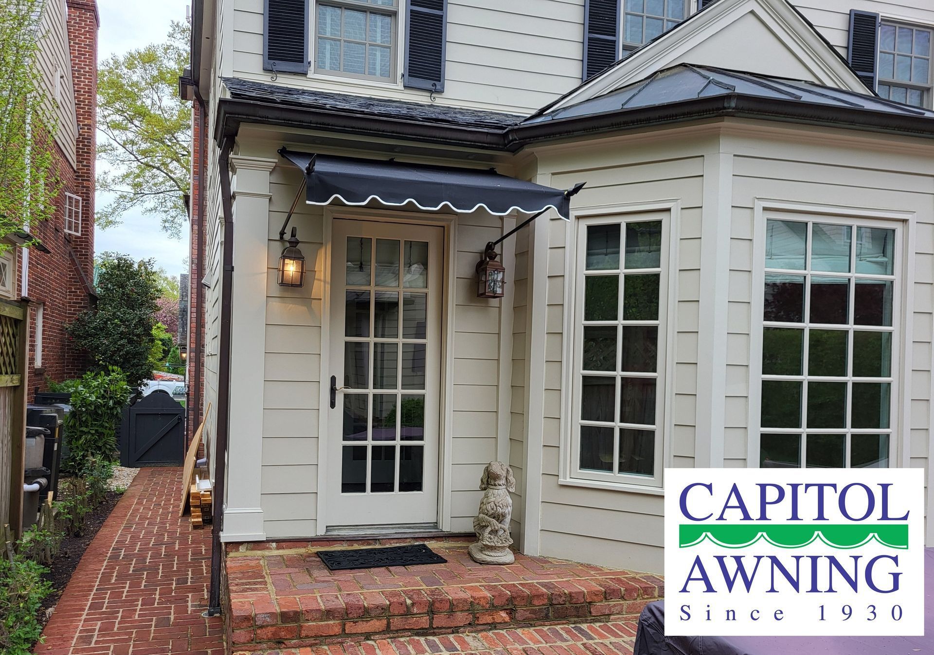 White house entry with glass door, lantern, brick path, and “Capitol Awning” logo in the corner