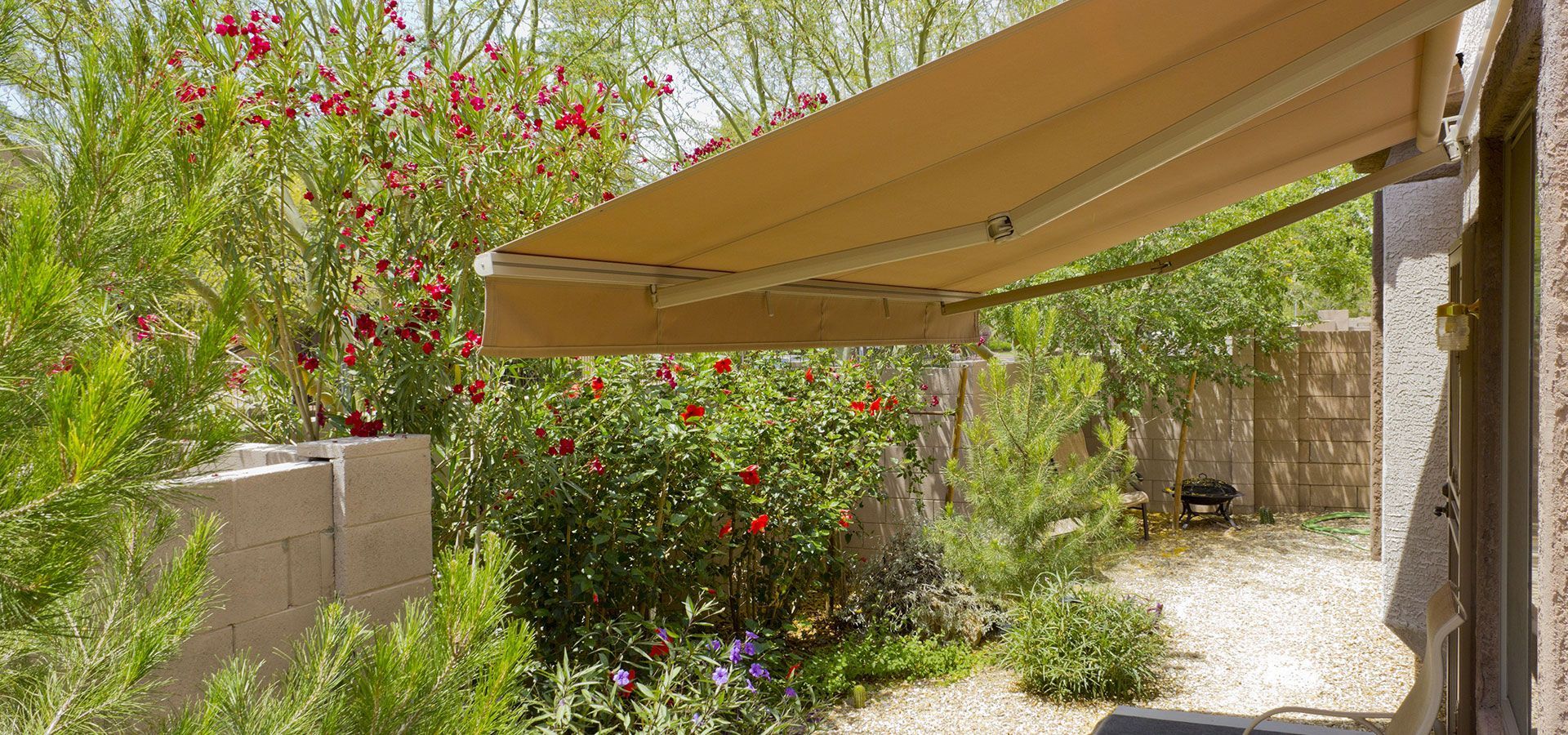 Patio with beige awning, stone wall, and lush flowering garden in bright sunlight