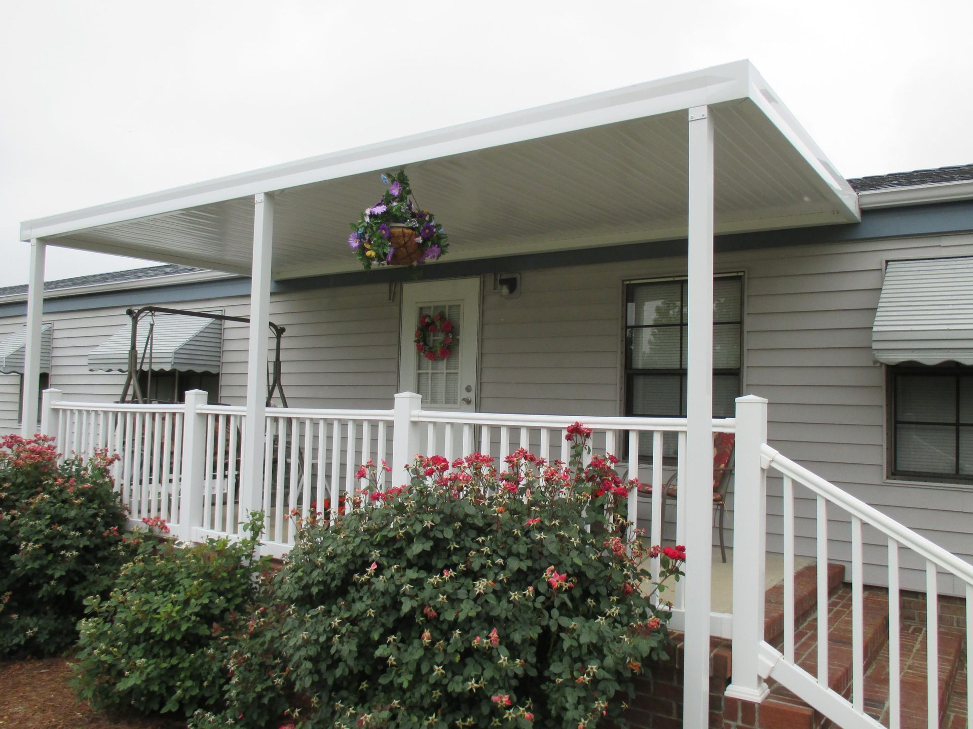 White porch with railings and steps, hanging flower baskets, and a large bush in front of a house.
