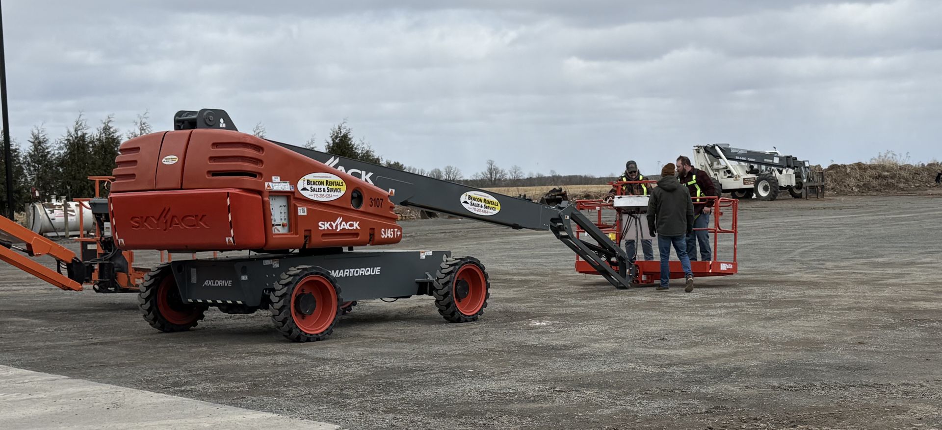 Red towable machine on gravel with two workers beside it under a cloudy sky.