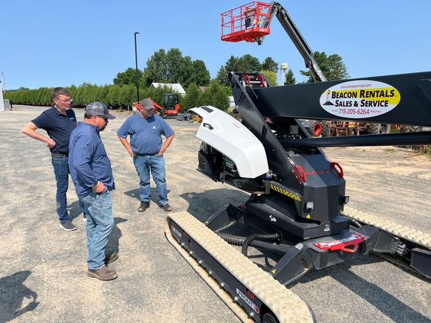 Three workers inspect a black and white aerial lift on a lot, with a red boom lift in the background.