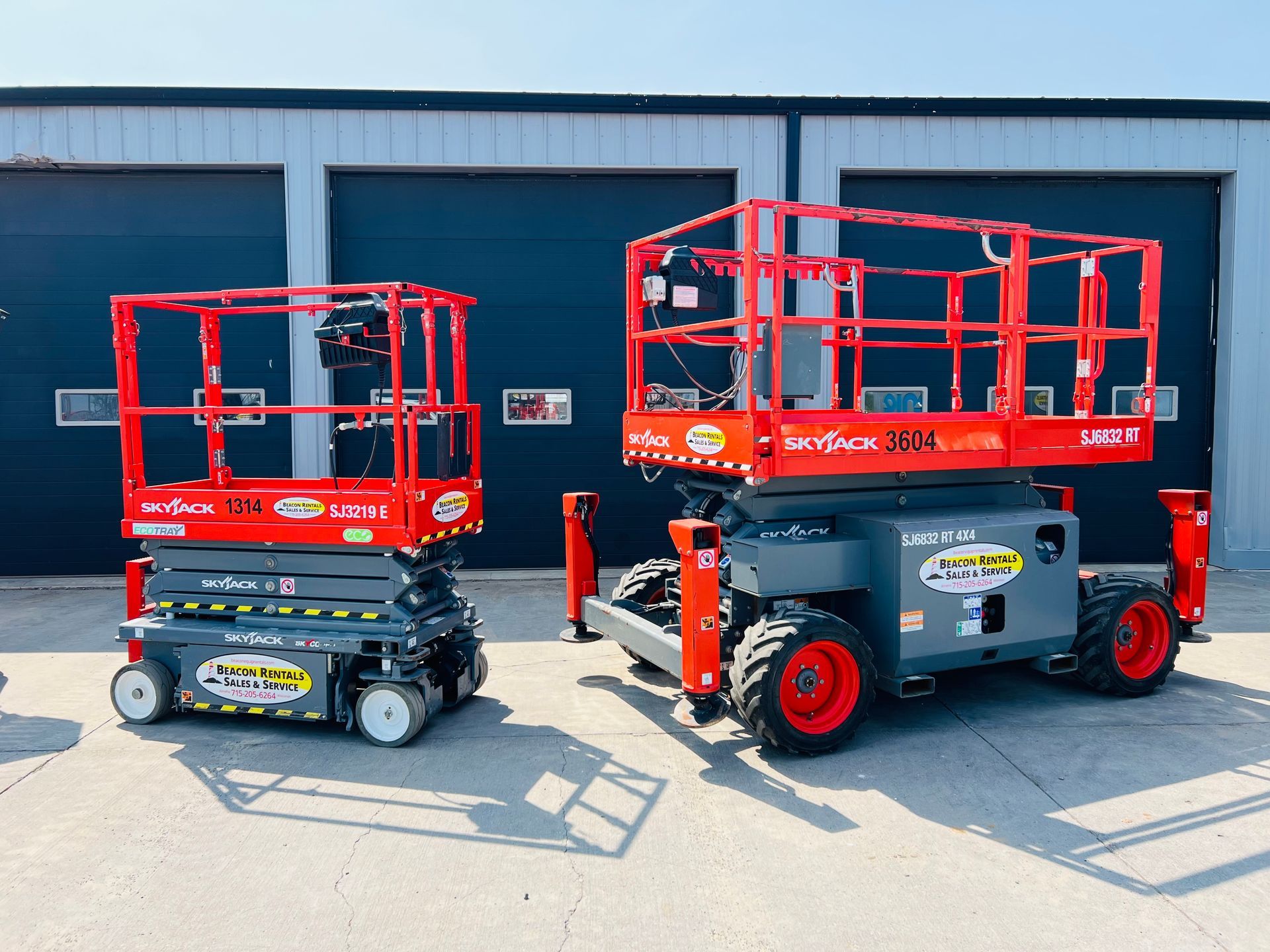 Two red scissor lifts are parked outside the garage doors on a concrete lot.