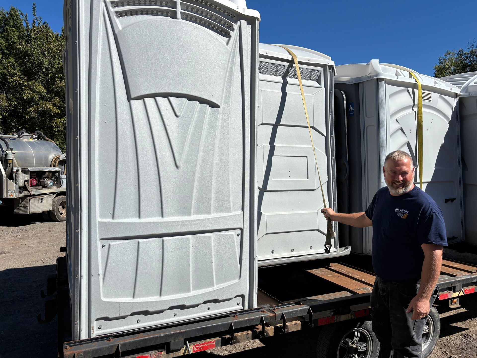 Portable toilets on a trailer, with a man standing beside them in a sunny outdoor lot