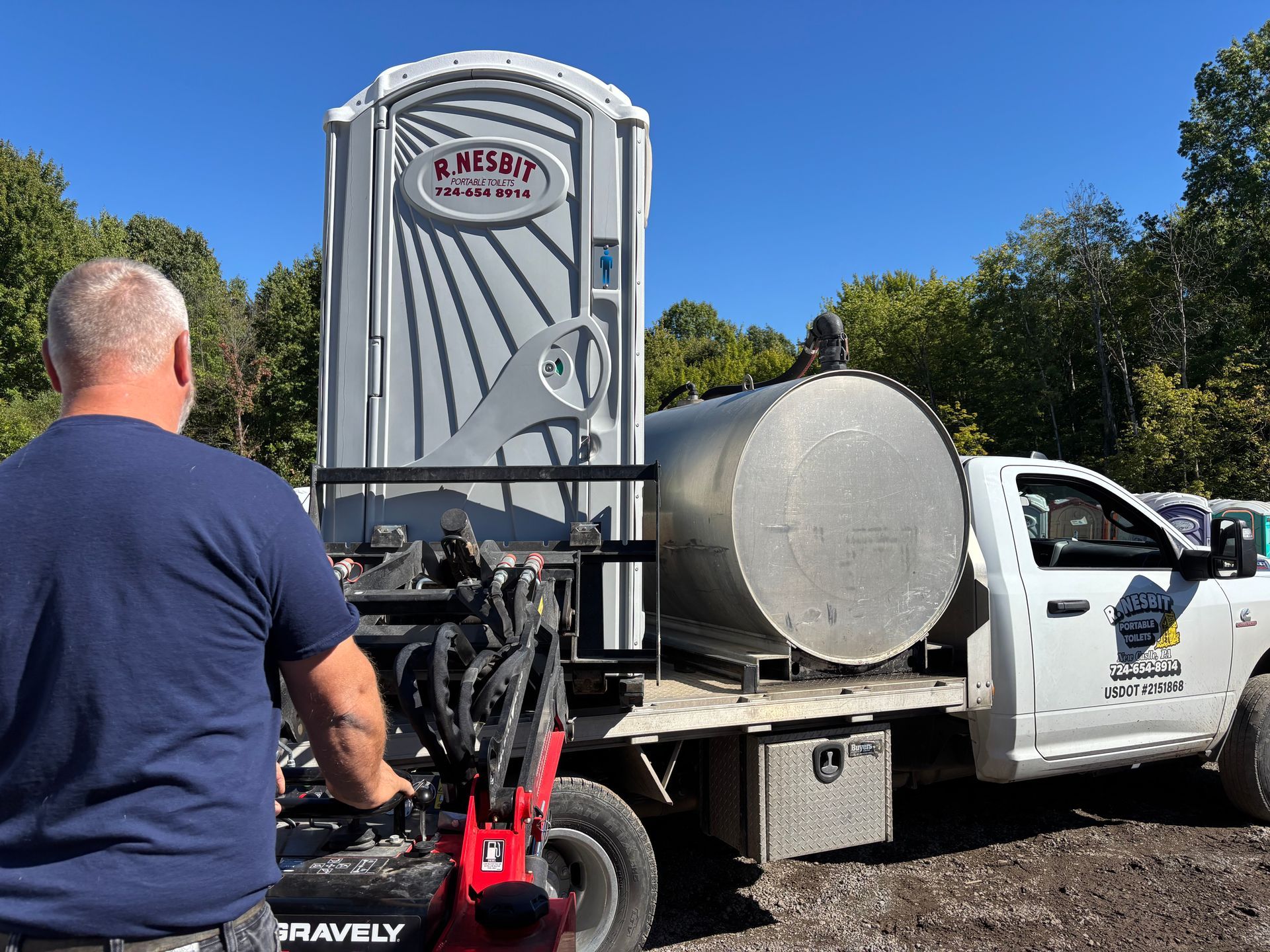 Worker beside a portable toilet on a utility truck in a sunny outdoor lot