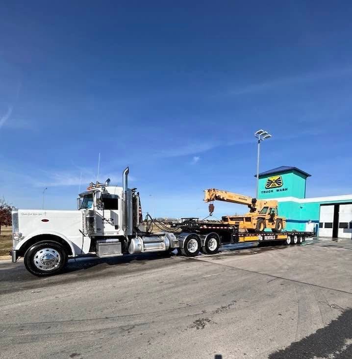 White semi-truck hauling a yellow crane, next to a teal and green building under a blue sky.