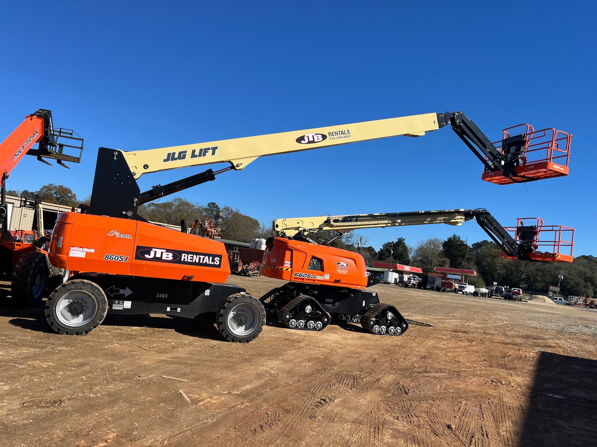 Two orange JLG boom lifts on dirt, one with wheels and one with tracks, under a blue sky.