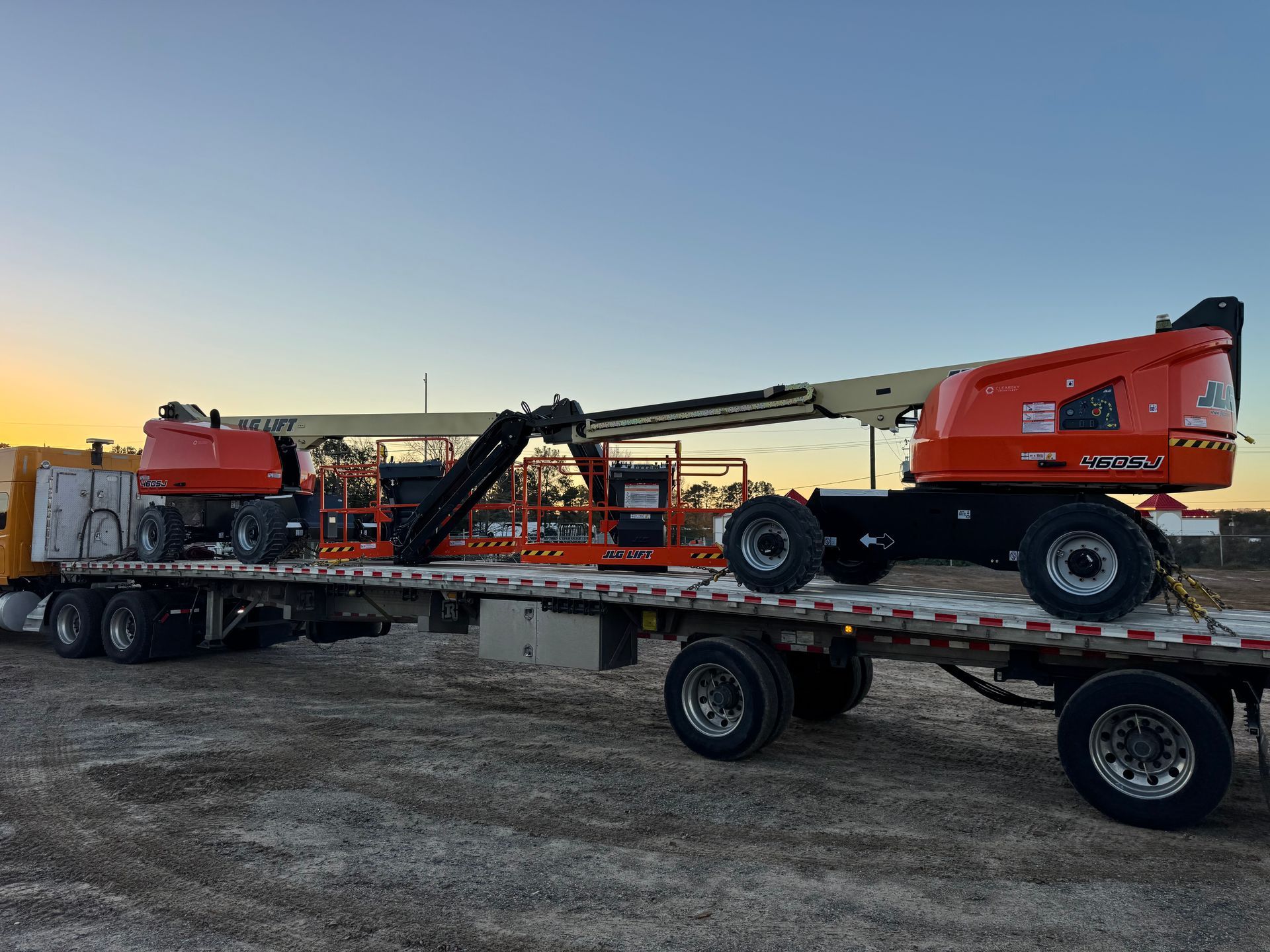 Two orange aerial lifts on a flatbed trailer, set against a sunset sky.