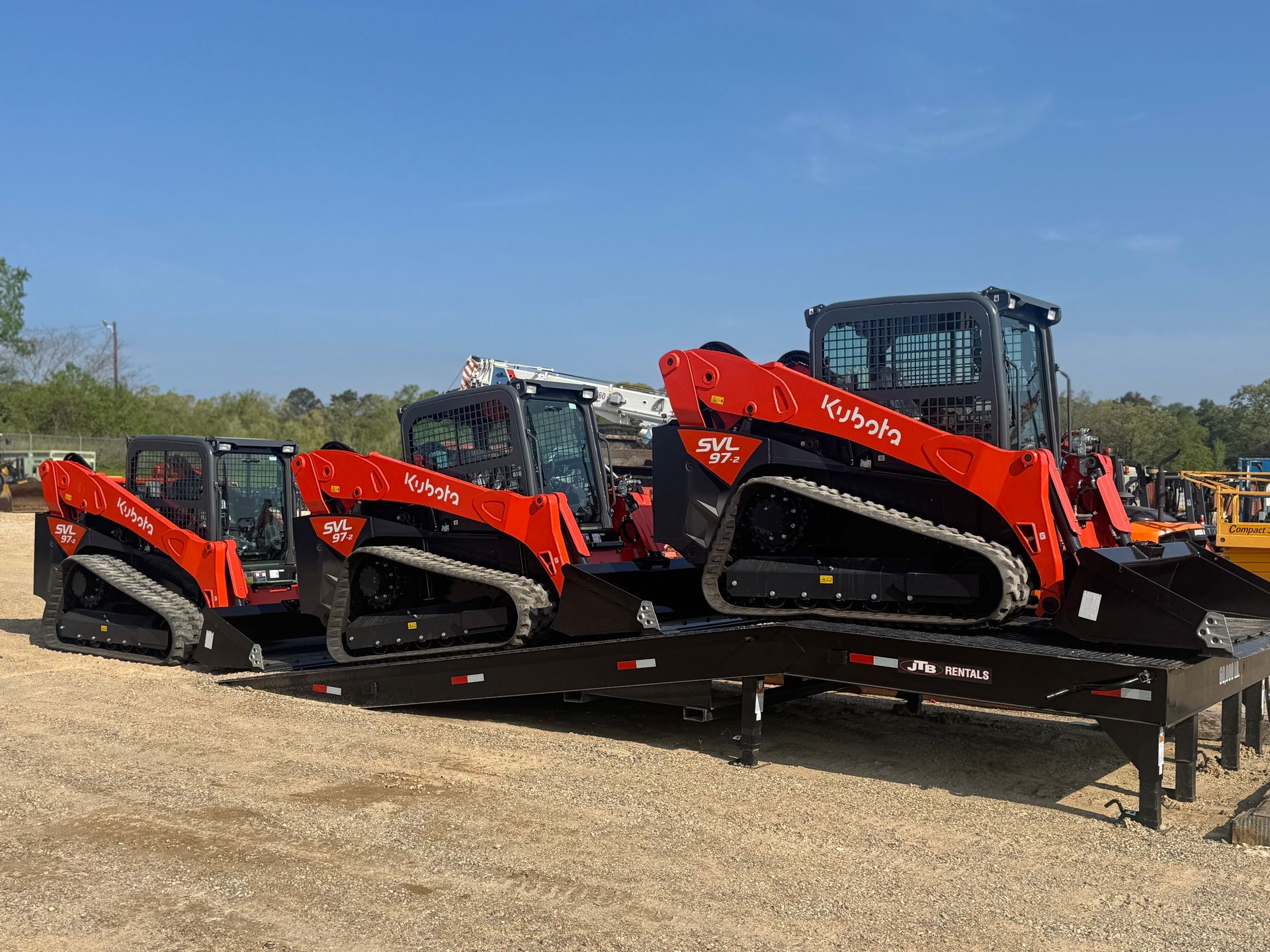 Three red and black track skid steer loaders on a flatbed trailer, outdoors under a blue sky.