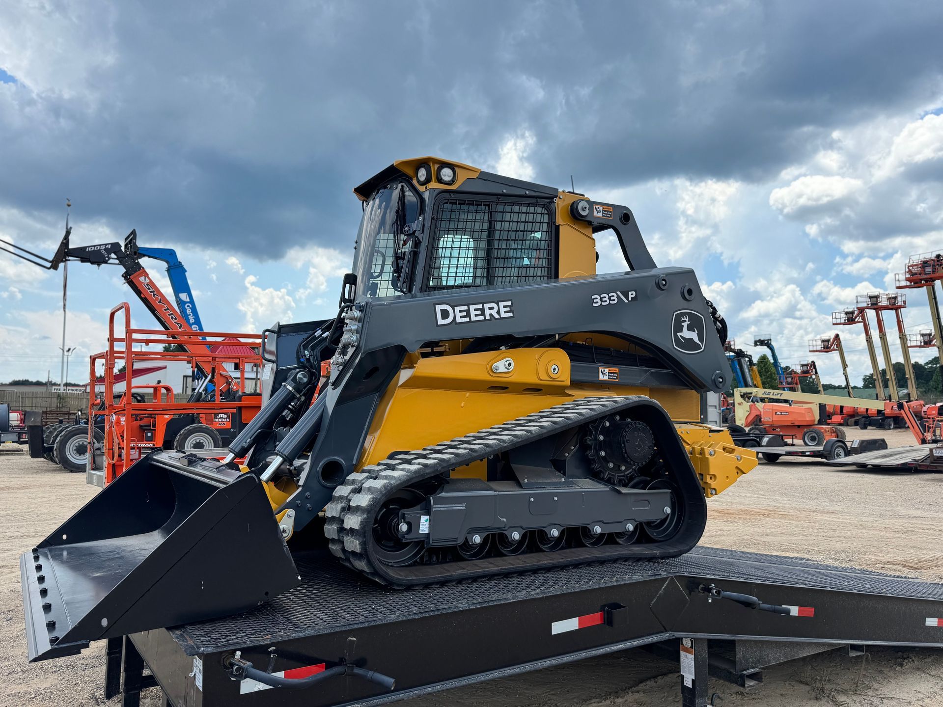 Yellow and black John Deere compact track loader on a trailer, under a cloudy sky.