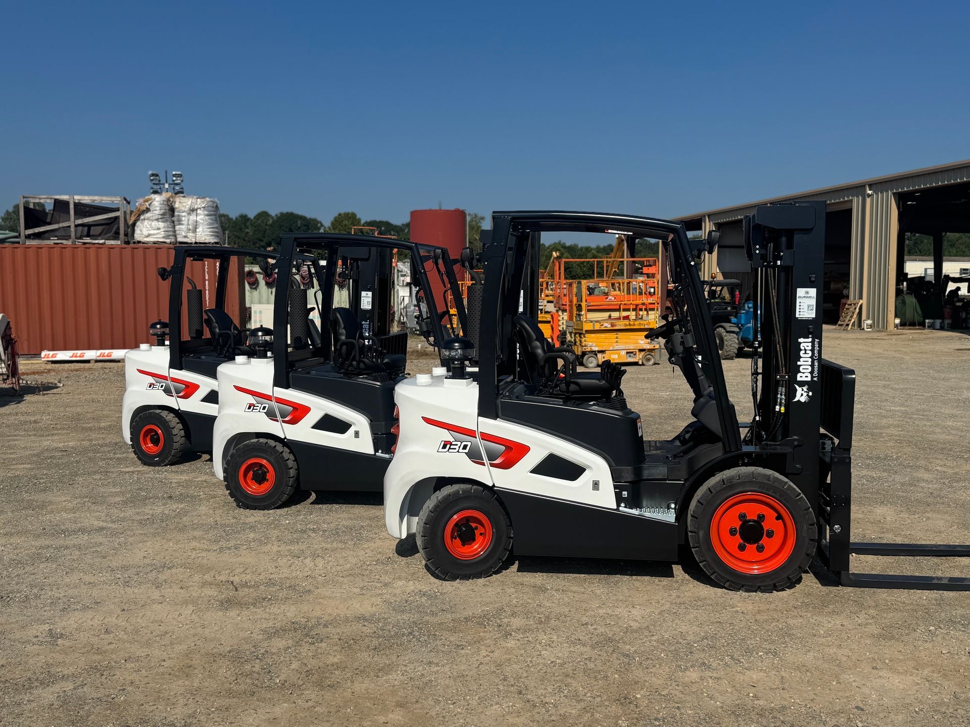 Three parked white and black forklifts with orange wheels on a gravel lot under a blue sky.
