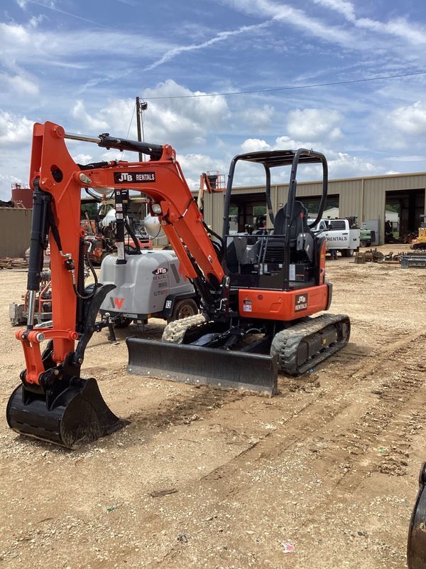 Orange and black mini excavator on gravel in front of a building on a sunny day.